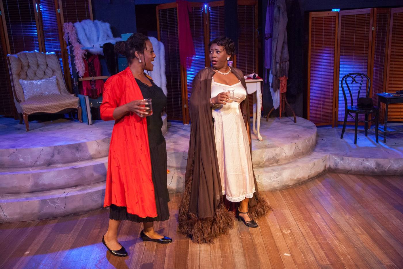 Production photo — two women in costume, red coat and feathered negligee