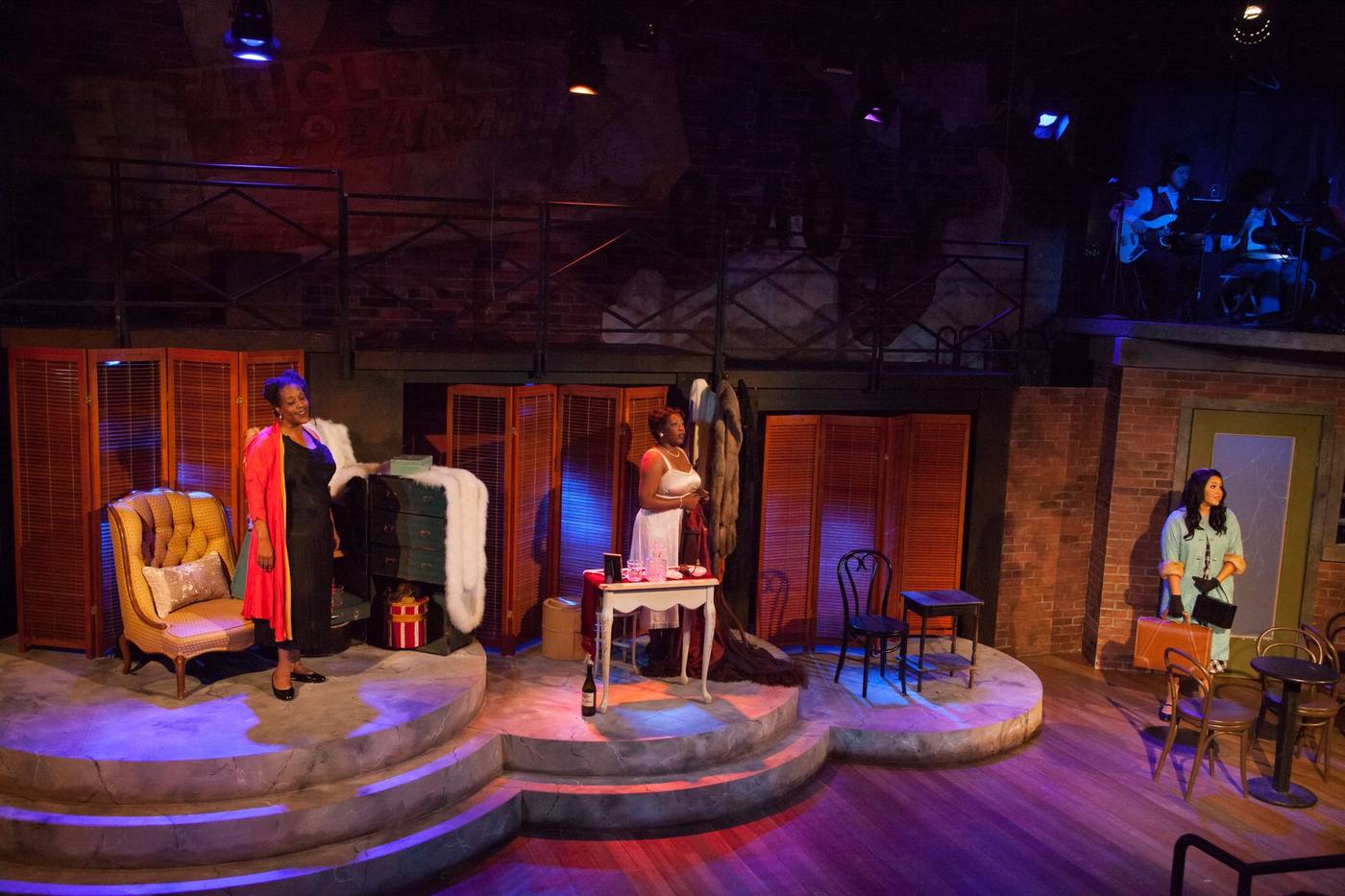 Production photo — three women on the marble-stepped set with louvred screens and catwalk