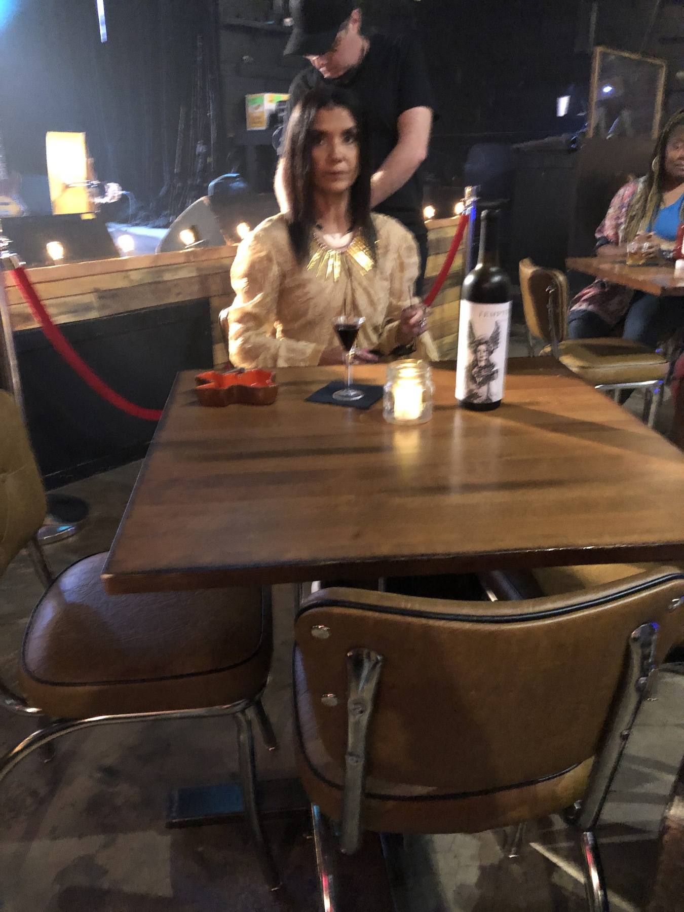 On set — lead female character in gold peasant blouse and statement necklace at bar table