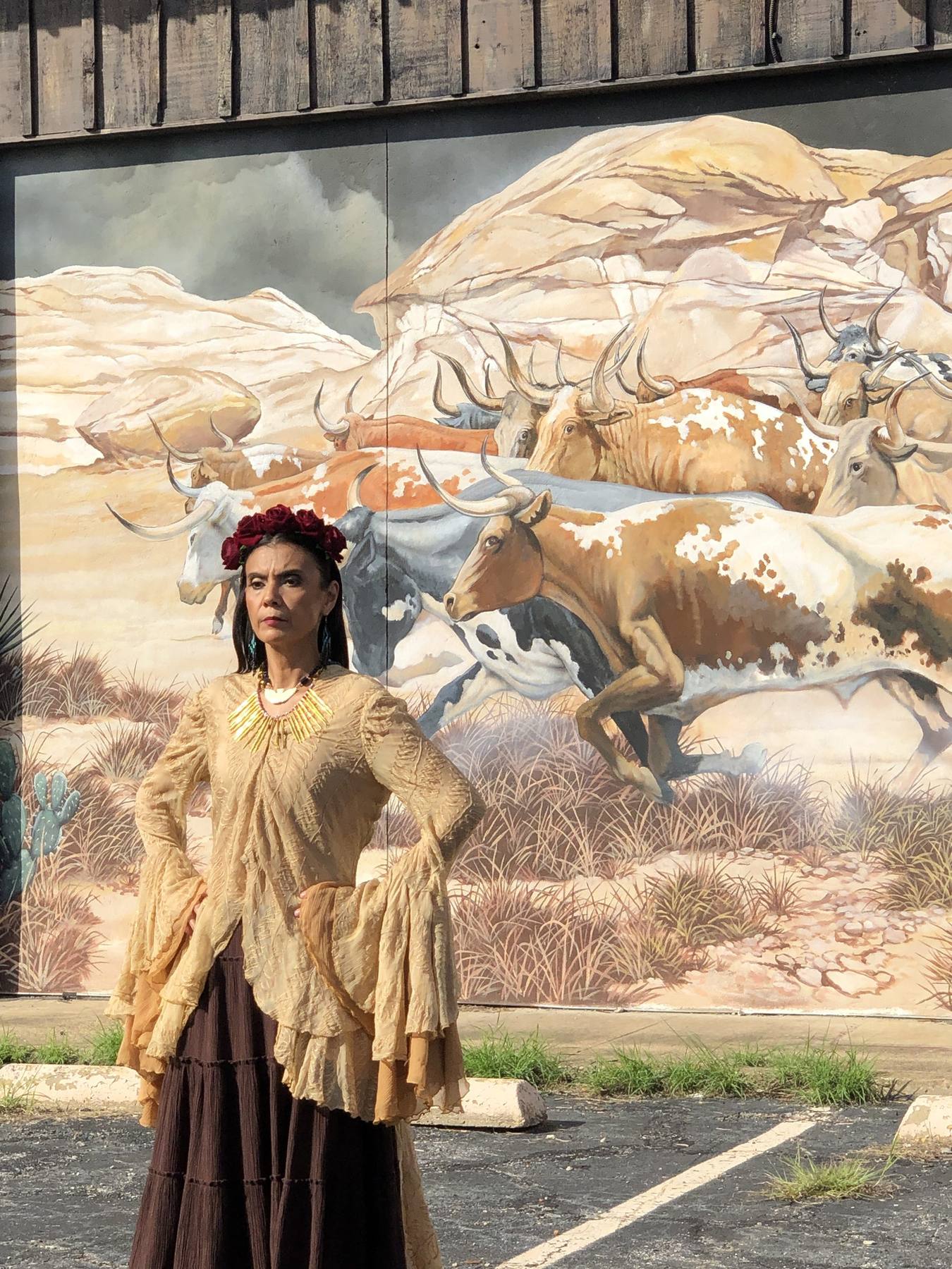 On set exterior — Frida-inspired character in golden blouse and tiered skirt against Texas longhorn mural