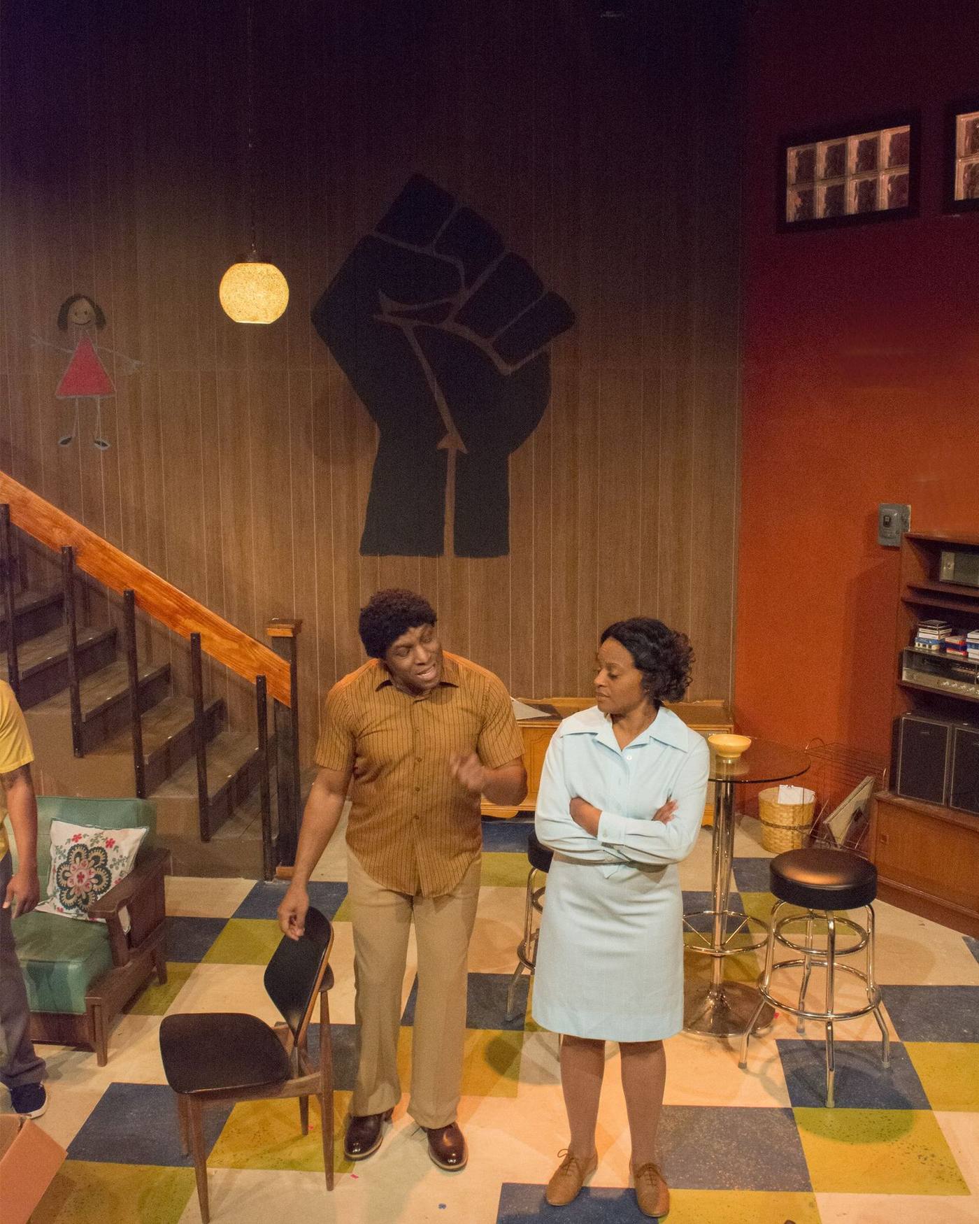 Production photo — confrontation with fist mural behind