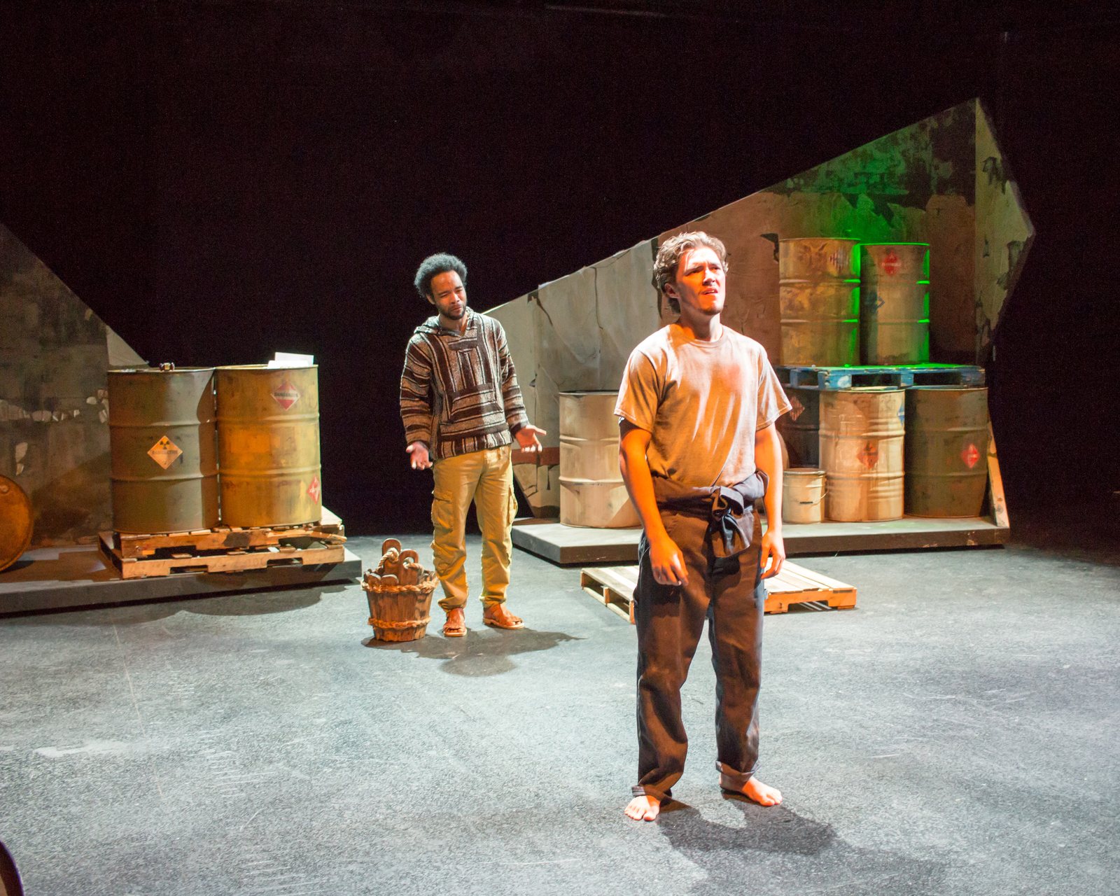 Production photo — two figures downstage among the drums