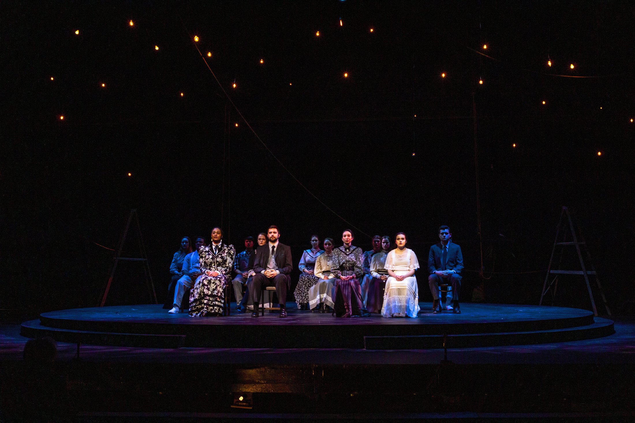Production photo — the company seated in a row under warm string lights