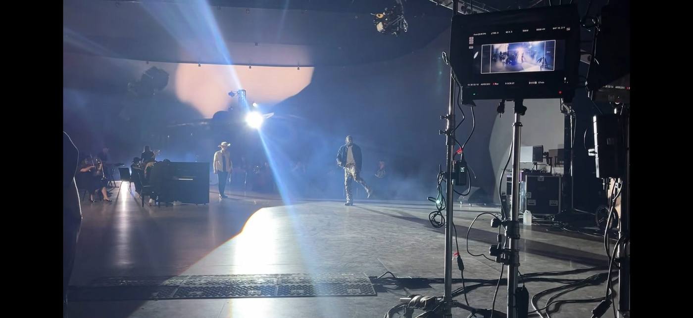 Camera monitor — both leads framed: country lead in cream western and cowboy hat, hip-hop lead in denim and printed pants