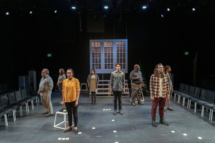 Playwrights in the Newsroom — the full company standing in the in-the-round newsroom set under the suspended window wall
