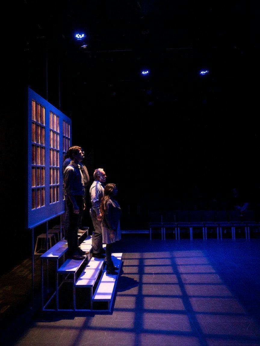 Production — an actor at the window wall lit in cold blue, panes glowing amber behind